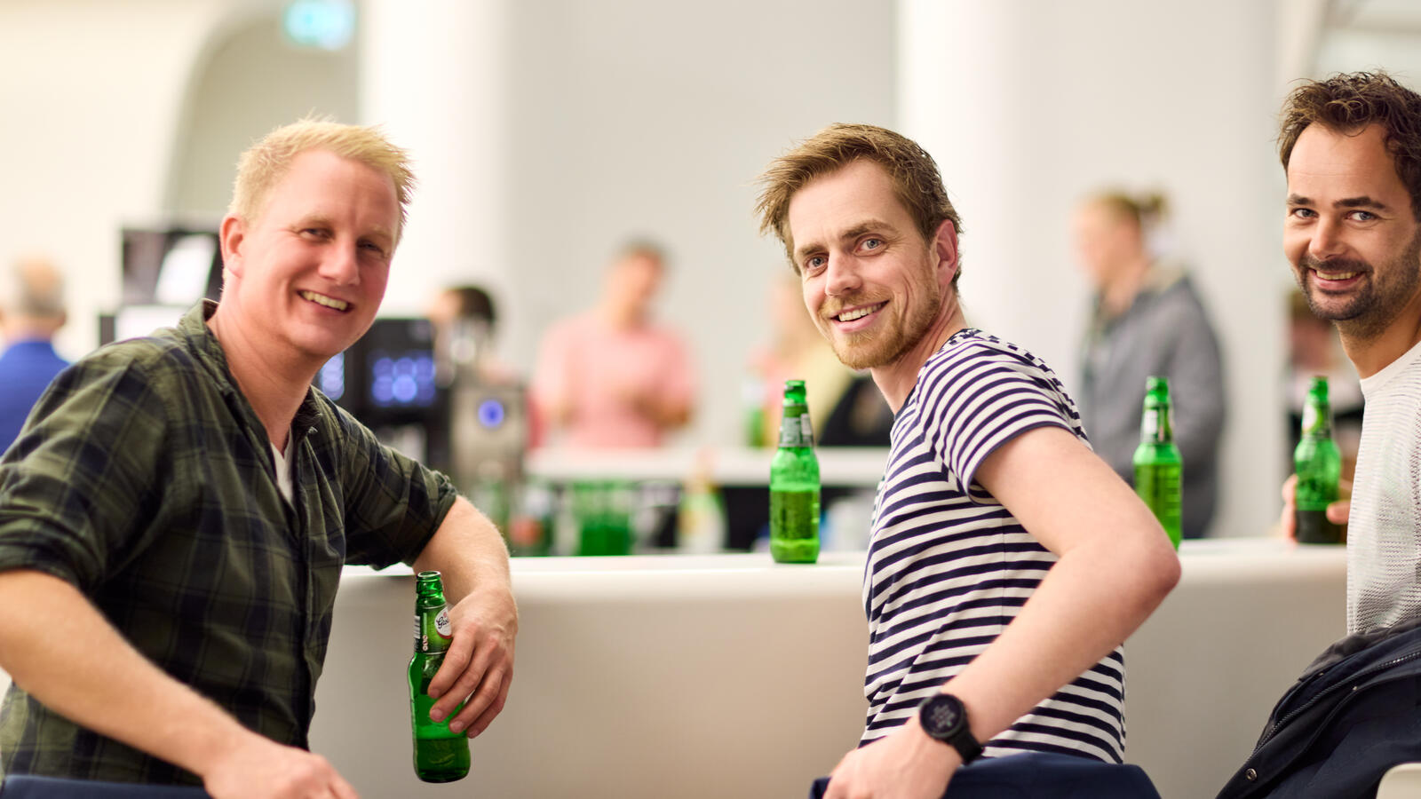 Drie jonge bezoekers zitten bij de bar van het museum met een biertje. Ze draaien zich om naar de camera en lachen.