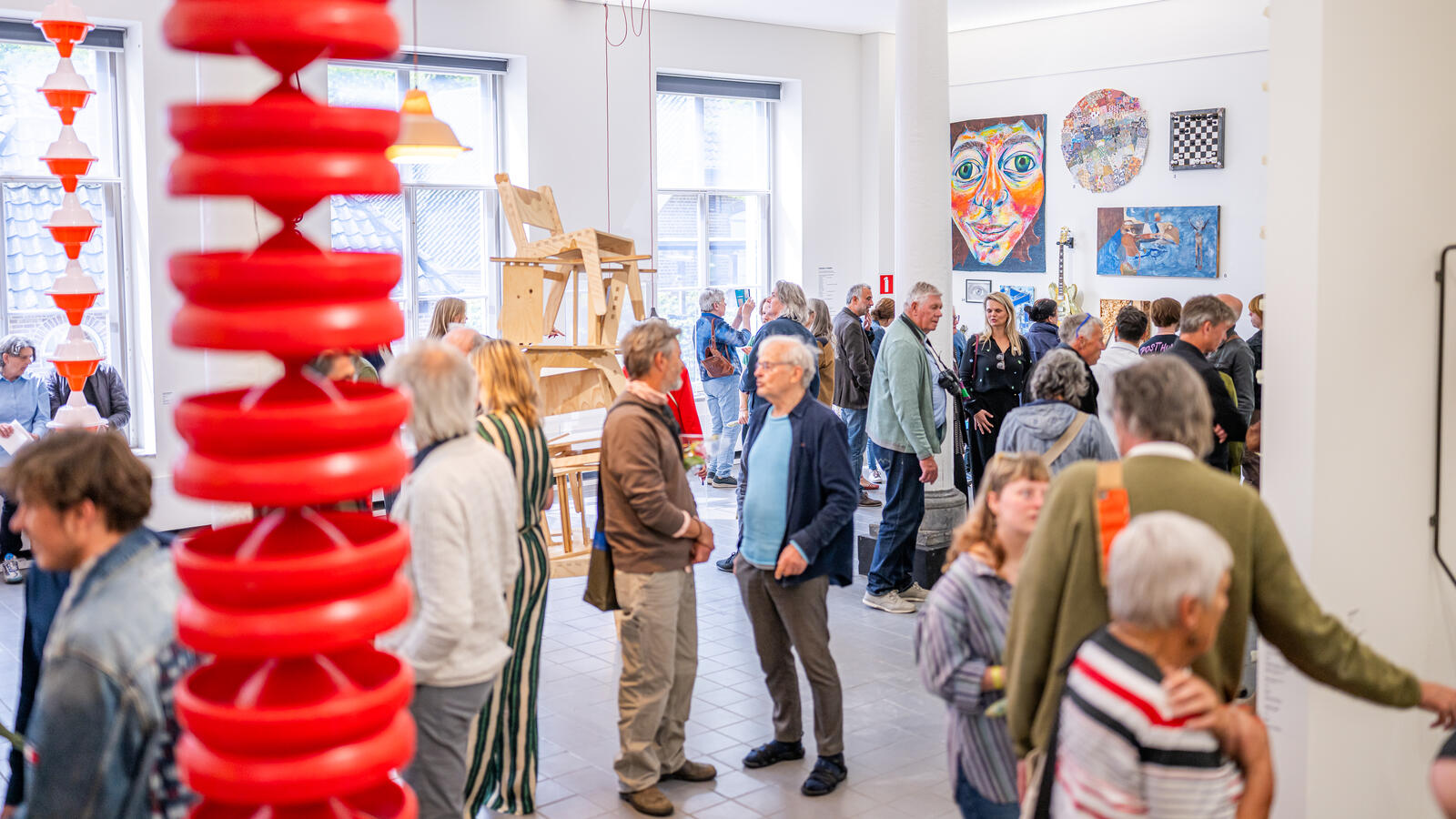 Tientallen bezoekers staan in de expositieruimte. Aan de voorkant van de foto hangt een rood kunstwerk van de plafond. 