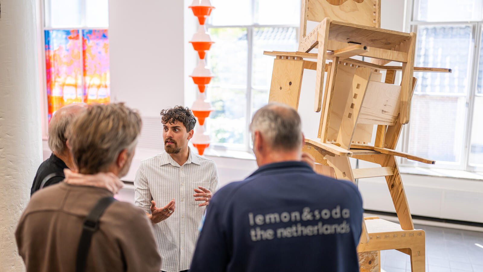 Een jonge kunstenaar staat bij een kunstwerk van op elkaar gestapelde houten stoelen. Hij spreekt een groep bezoekers toe.
