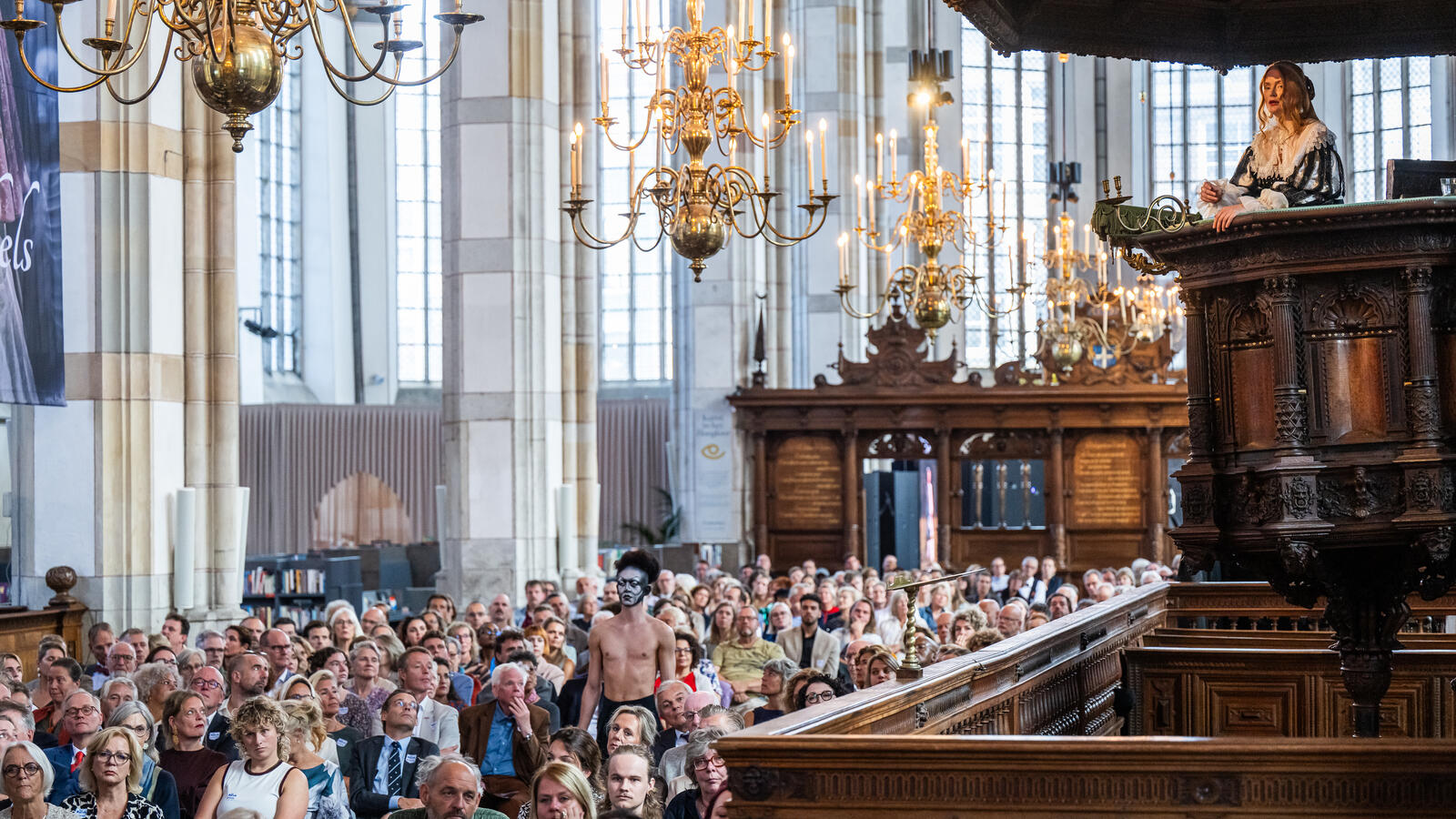 In de Grote Kerk staat een actrice in 17e eeuwse kleding op het houten spreekstoel. Honderden mensen zitten in de zaal en kijken naar haar. Boven hen hangen kroonluchters.