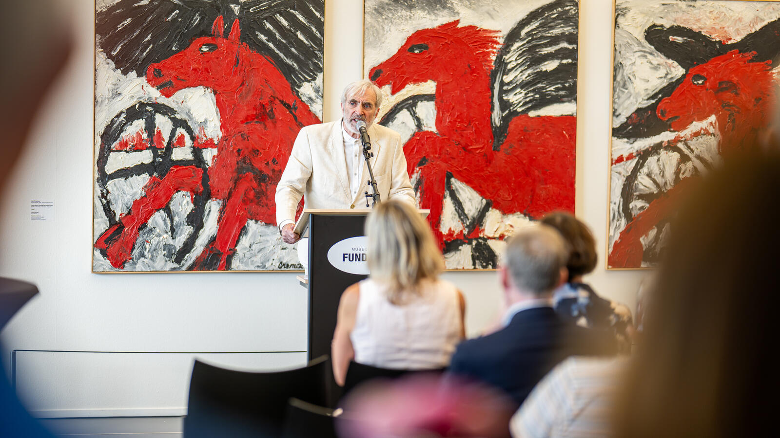 Henk Heideveld staat voor grote schilderijen van rode paarden en spreekt het publiek toe.