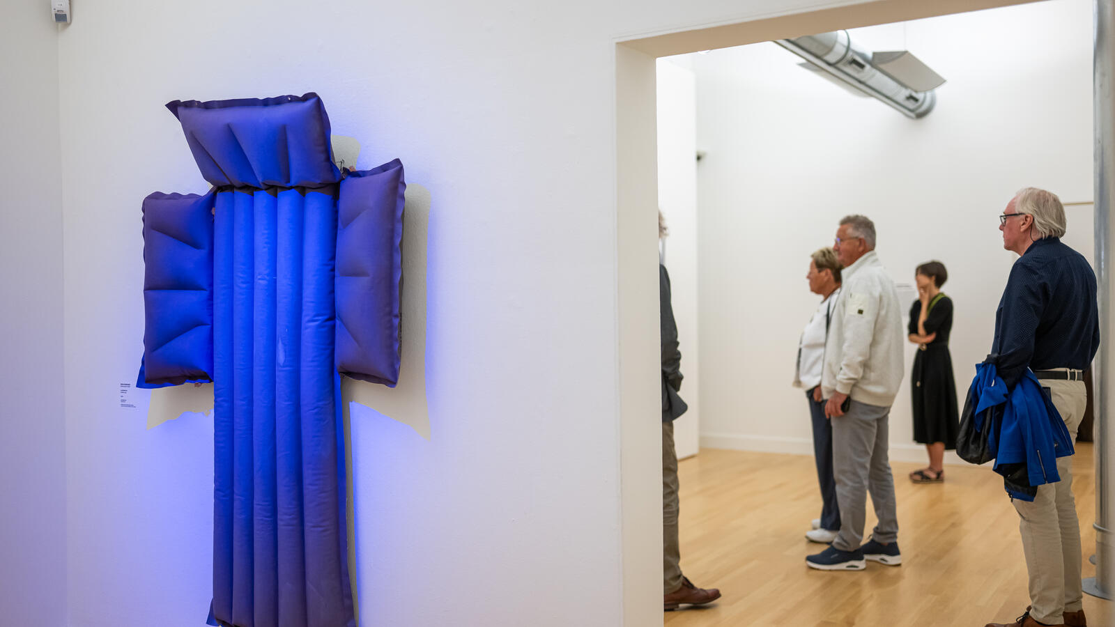 Aan de muur hangt een blauwe kunstwerk in de vorm van een kruis dat lijkt op een luchtbed.