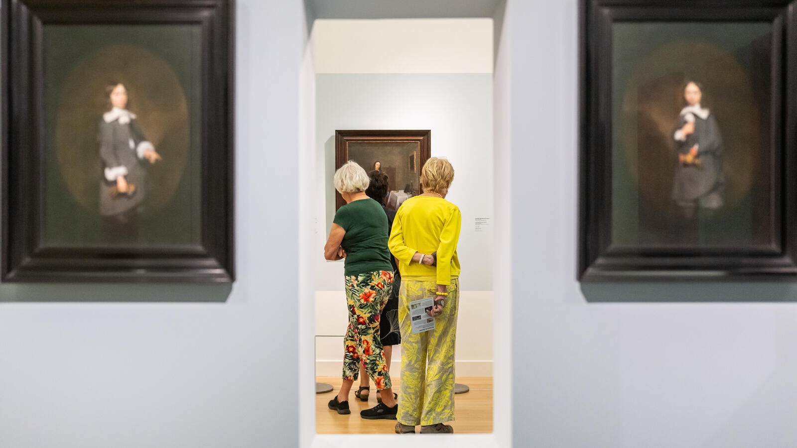 Twee bezoekers staan voor een schilderij. De foto is gemaakt door een doorkijkje. Links en rechts van het doorkijkje hangen twee portretten van Ter Borch.