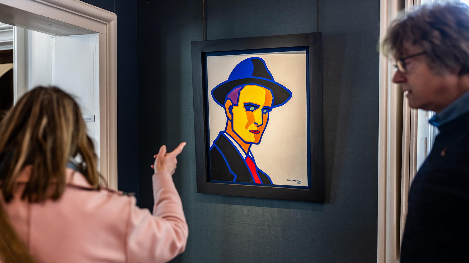 Een bezoeker in een roze jas wijst naar een schilderij van een man met een hoed op die schuin naar haar lijkt terug te staren.
