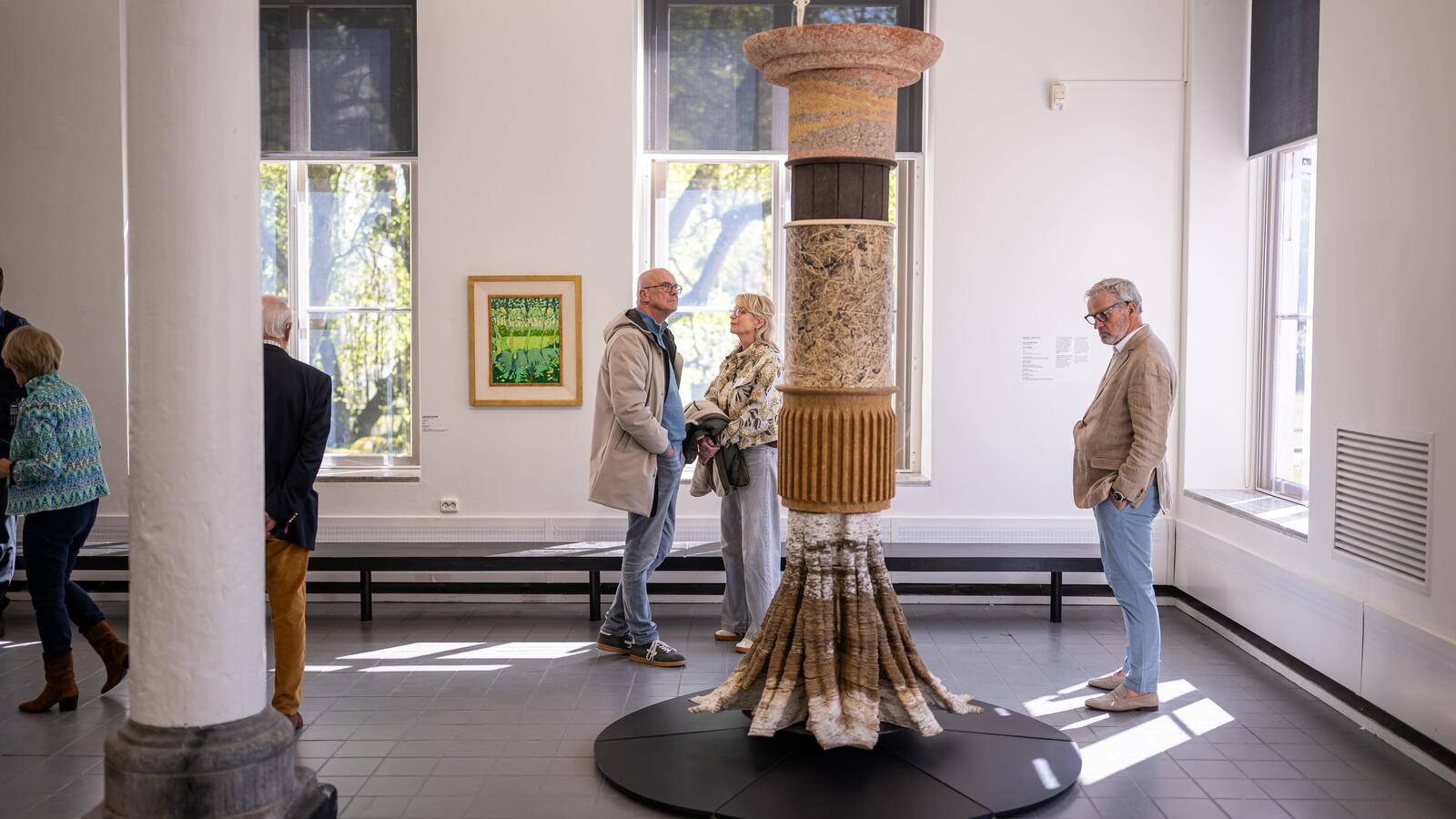 Een ruimte met grijze vloer en witte muren. In het midden staat een kunstwerk: een pilaar van verschillende materialen. Bezoekers kijken ernaar.