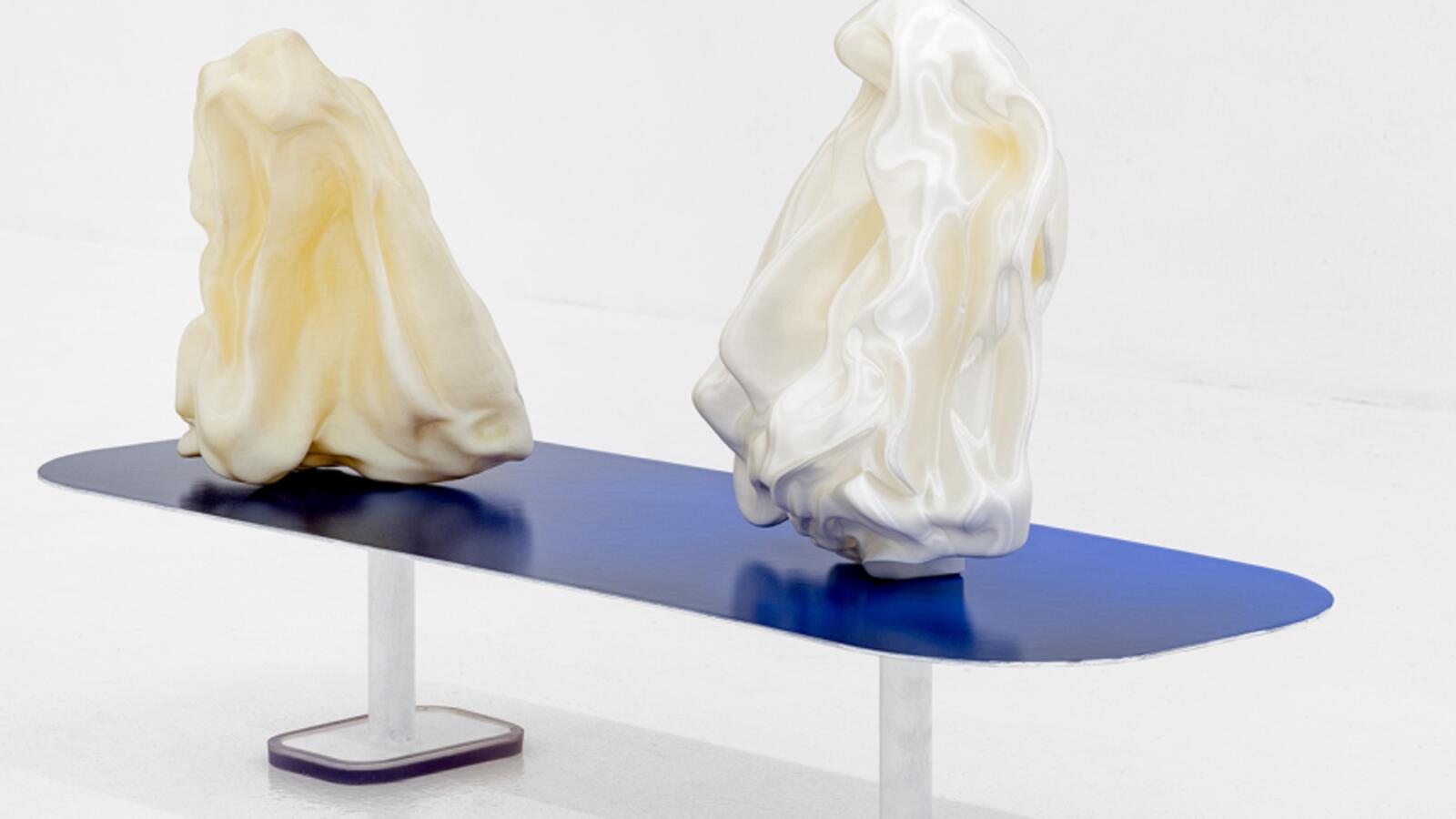 Twee witte sculpturen die lijken op proppen papier of zijde, rustend op een blauwe tafel.