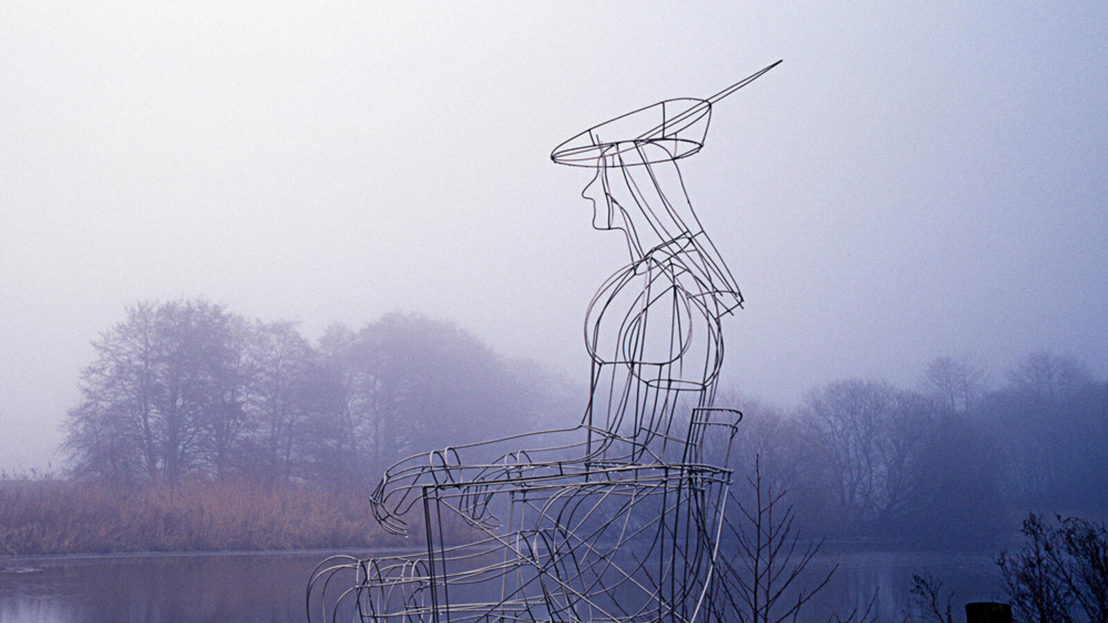 Een ijzeren draadsculptuur van een zittende menselijke figuur staat aan het water, bijna doorschijnend tegen een mistig landschap met bomen en een blauwgrijze lucht.