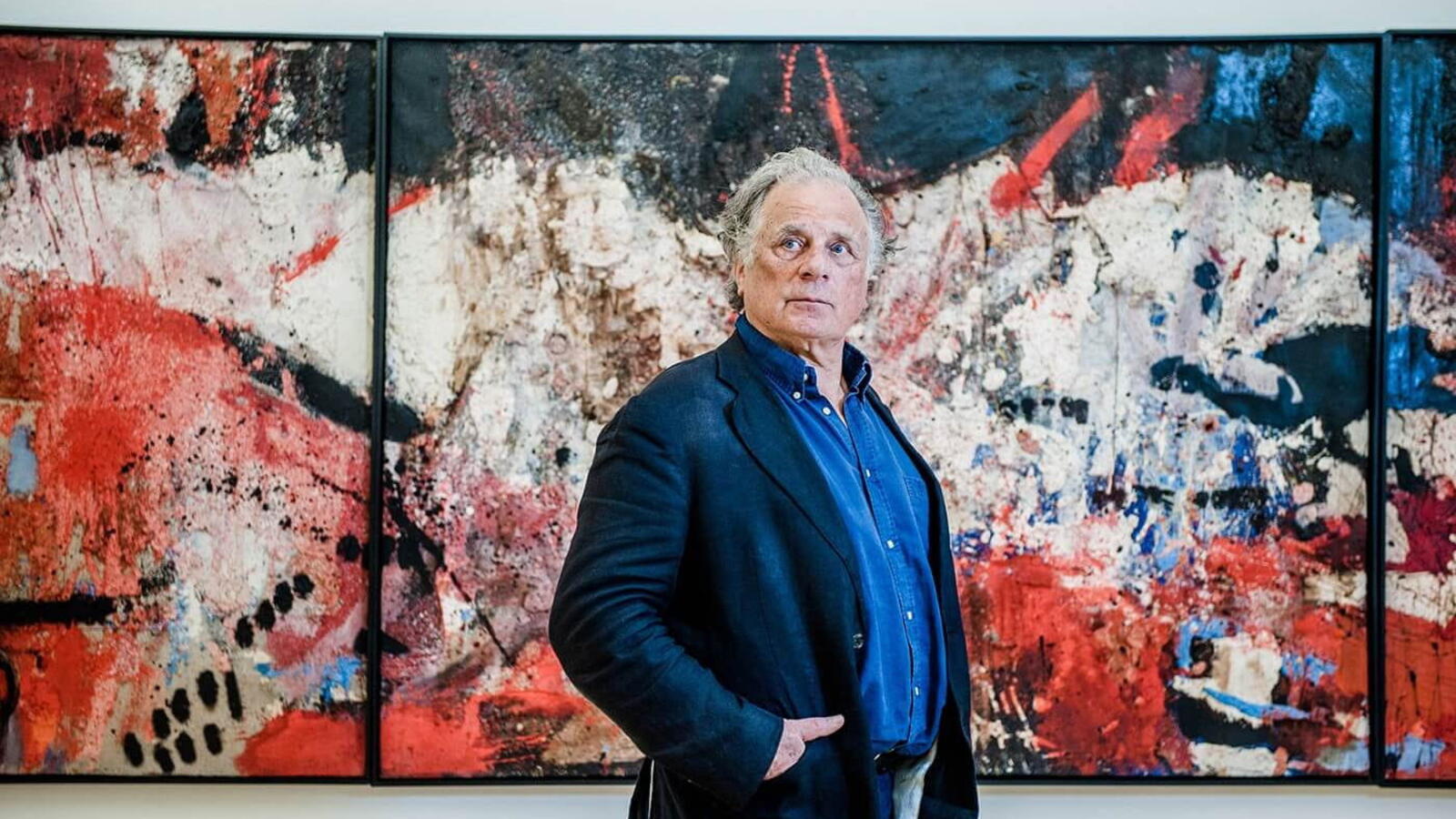 Een oudere Jan Cremer staat voor een groot schilderij in rood en donkerblauw. Cremer draagt zelf een donkerblauw pak en kijkt naar de verte.
