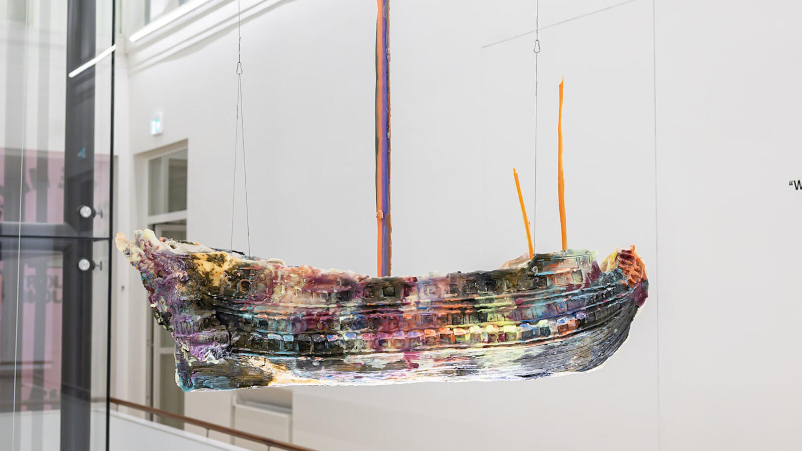 Kleurrijk, bootvormig kunstwerk hangt met dunne draden in een lichte museumruimte. Op de achtergrond zijn witte muren, een trapleuning en glazen delen van het gebouw te zien.