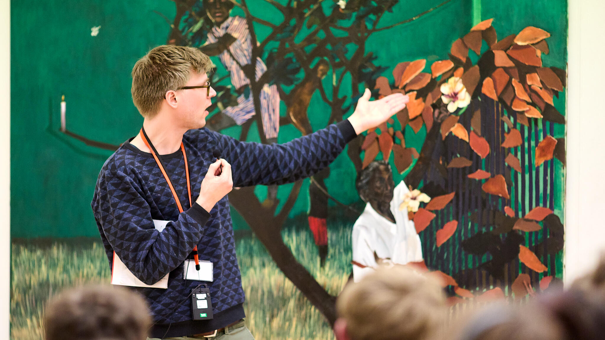 A male museum docent stands in front of a green painting. He is pointing to the right and talking. In the foreground, you can see the backs of listening students.