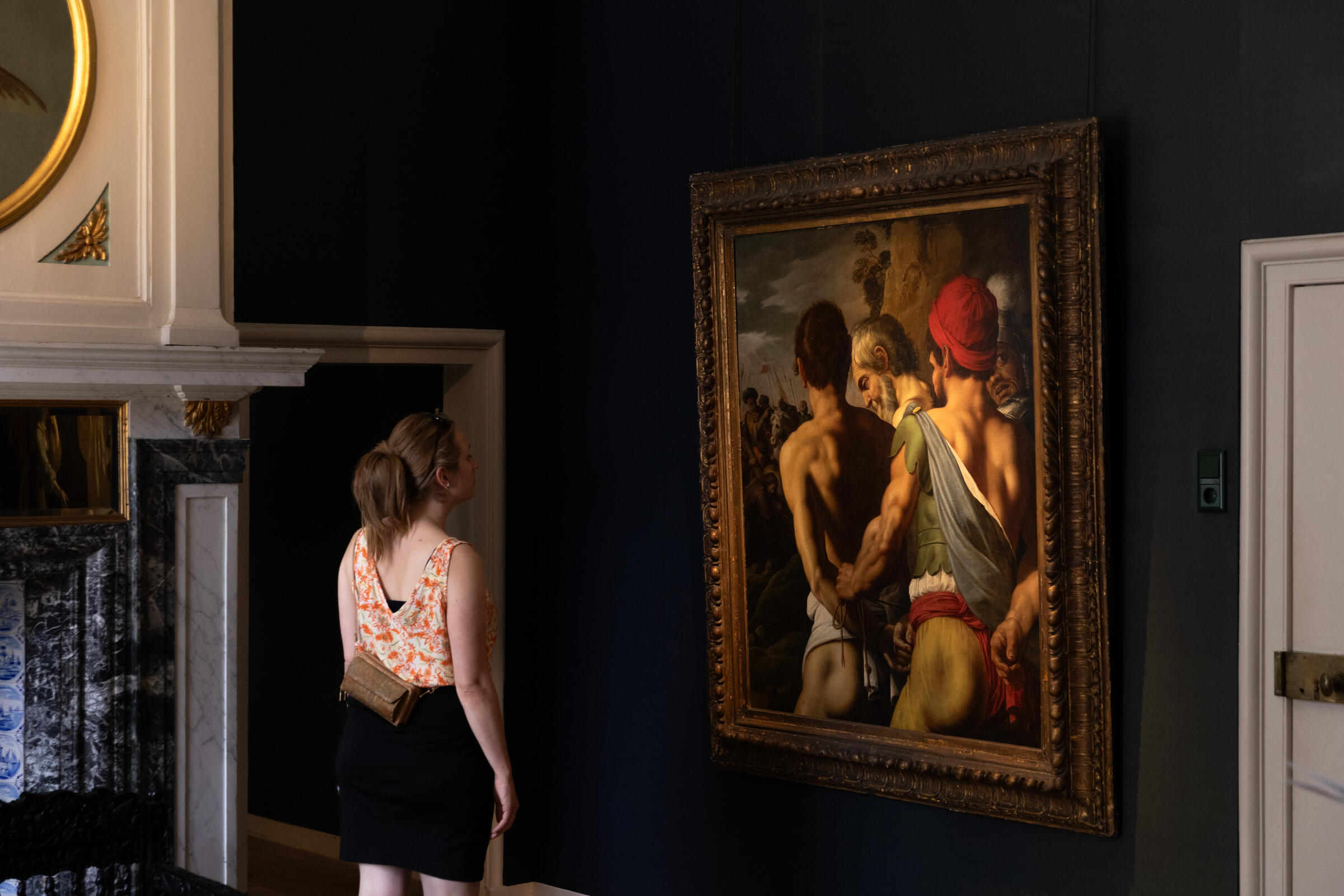 Een jonge vrouw staat voor een groot schilderij van twee mannen in kasteel het Nijenhuis. De ruimte is donkerblauw met statige witte accenten. Het schilderij hangt in een grote goudkleurige lijst en wordt mooi belicht door een spotje.