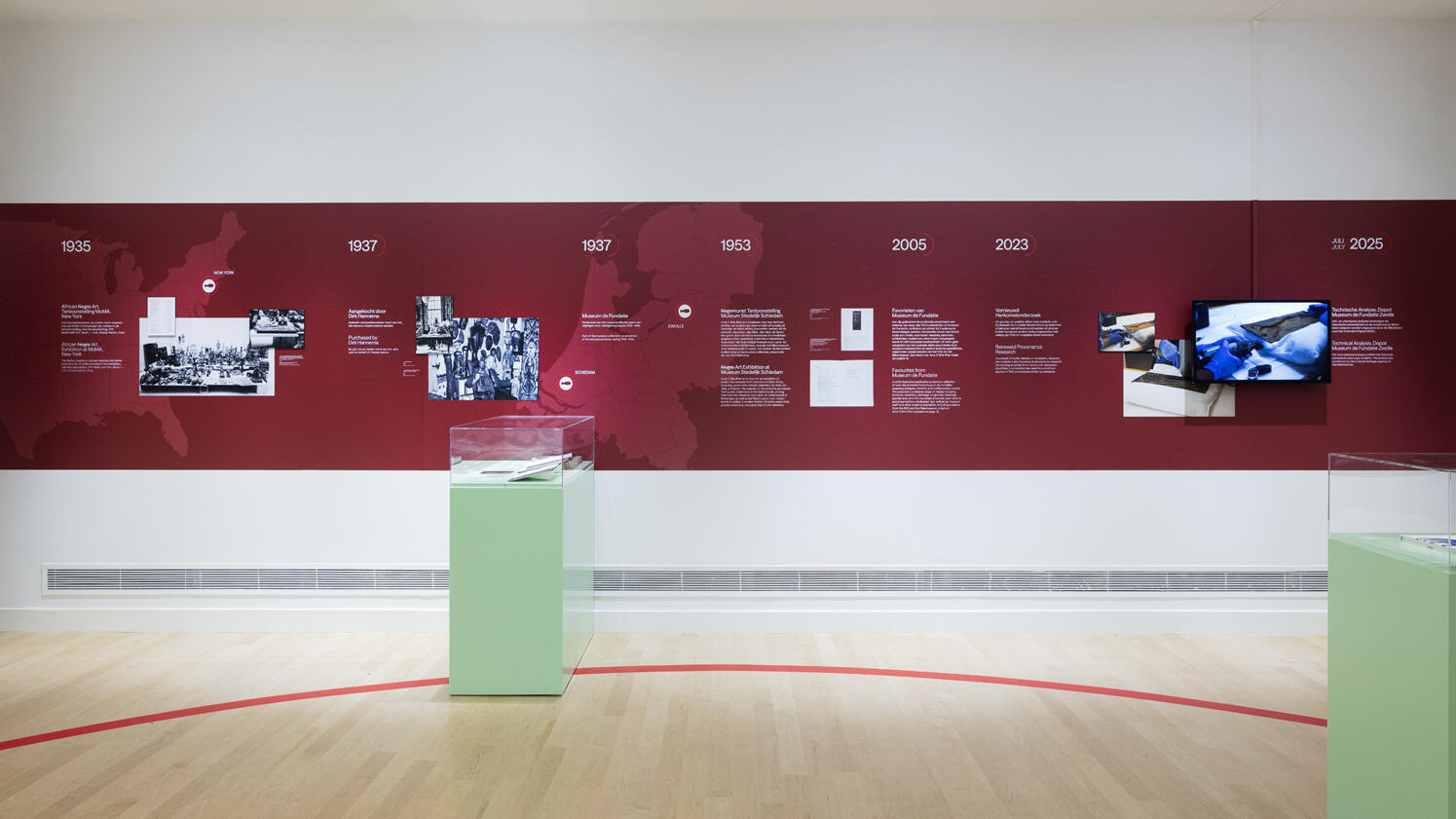 Een muur van het museum met daarop een rode strook op de muur. Op deze strook staat allemaal informatie en foto's van het herkomstonderzoek van Back to Benin.
