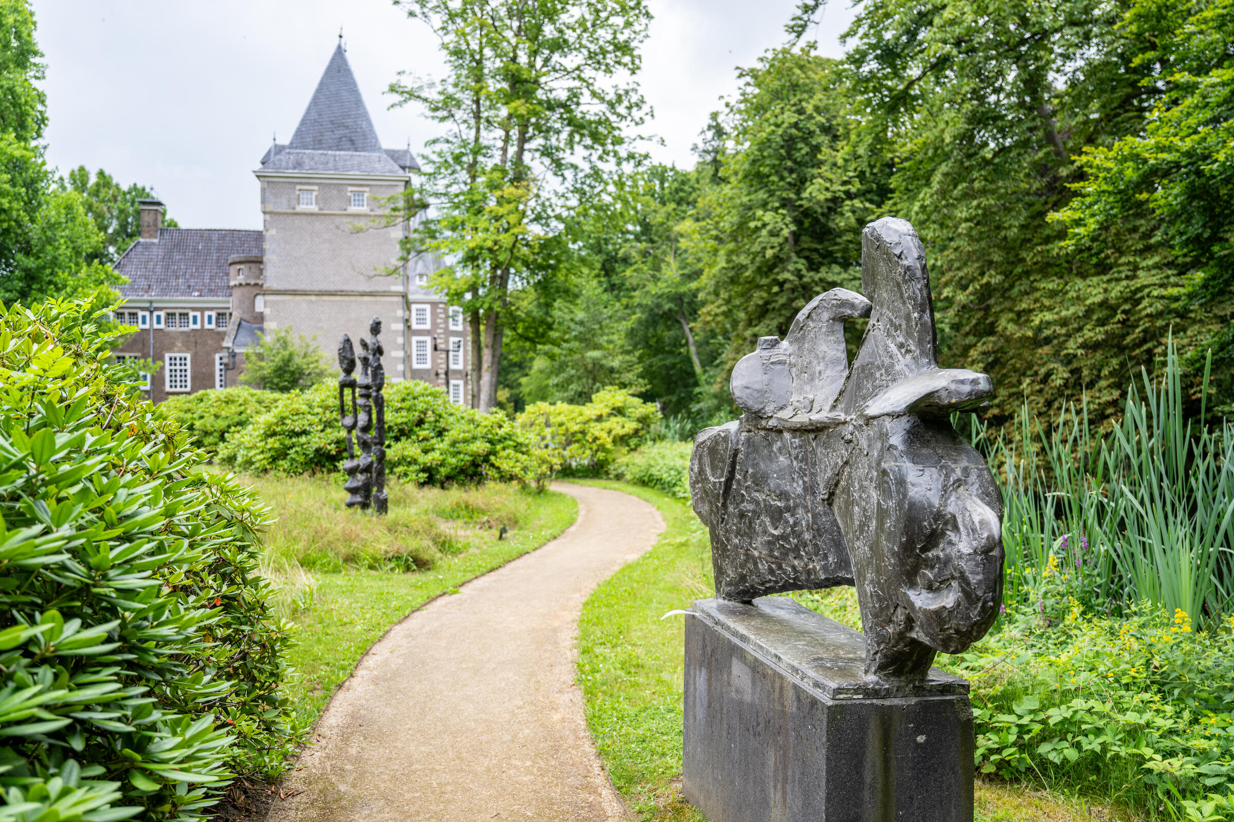 Een stenen sculptuur staat op de voorgrond. Een kronkelende weg loopt in de verte naar het kasteel, omringd met groen.
