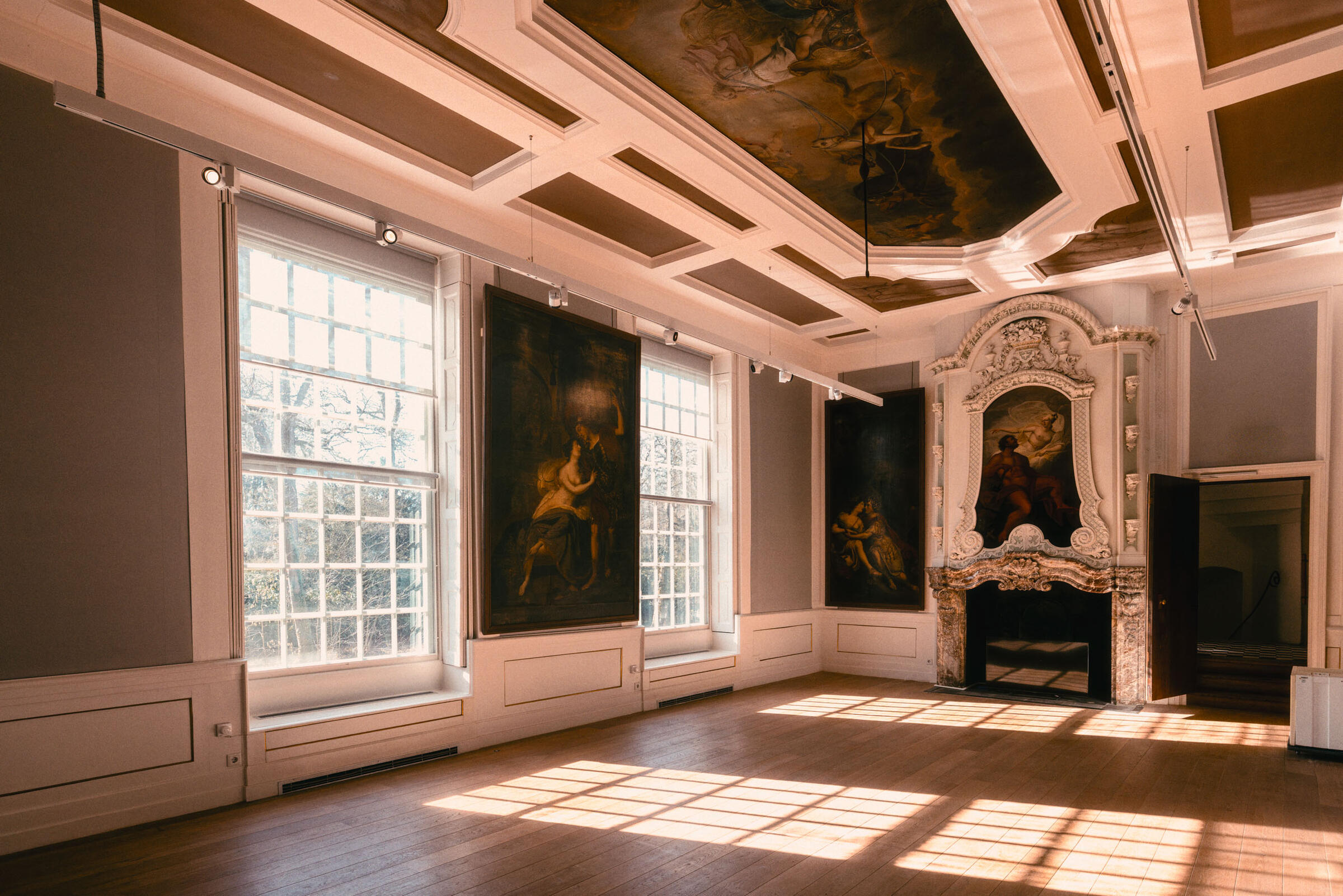 Een statige zaal in Kasteel het Nijenhuis met kunst aan de muren en op het plafond. Het zonlicht schijnt naar binnen door de grote ramen.