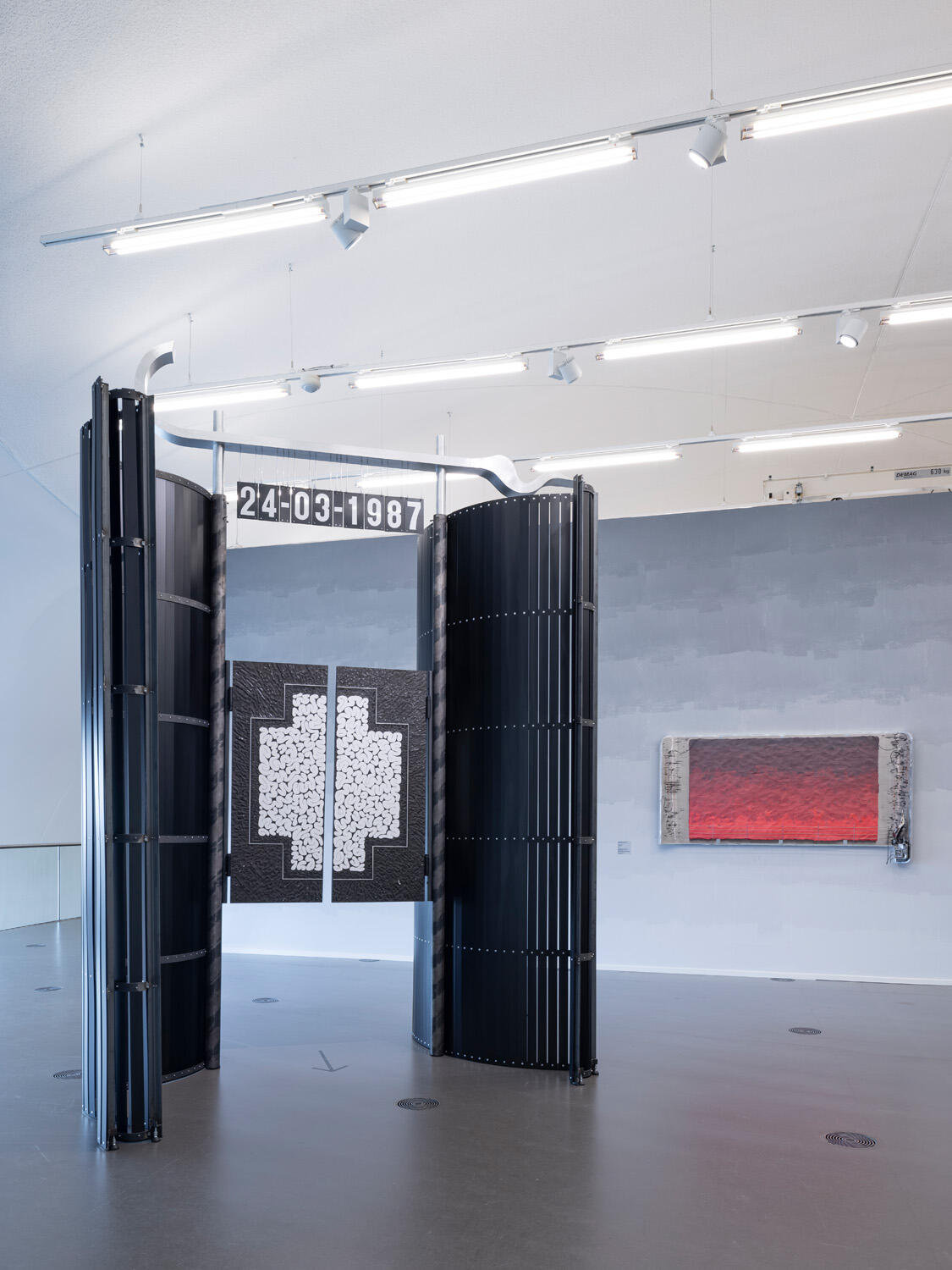 Een ruime museumzaal met links een grote zwarte installatie van gebogen panelen. In het midden hangt een zwart-wit kruisvormig kunstwerk met daarboven de datum 24-03-1987. Rechts aan de muur hangt een langwerpig kunstwerk met een rode gloed.