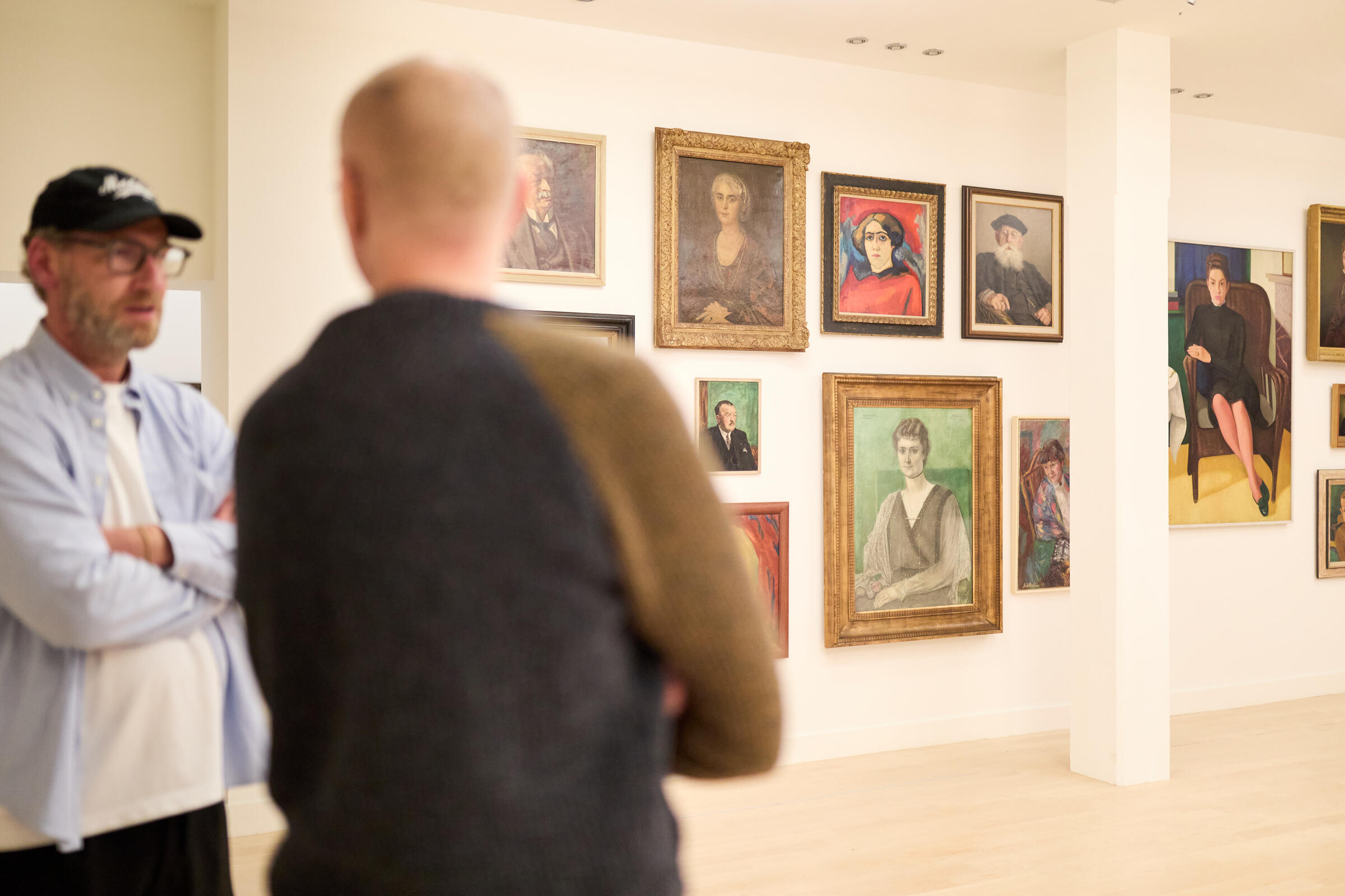 Twee mannelijke bezoekers praten met elkaar in een zaal waar allemaal portretten van vrouwen aan de muur hangen. De mannen zijn onscherp.