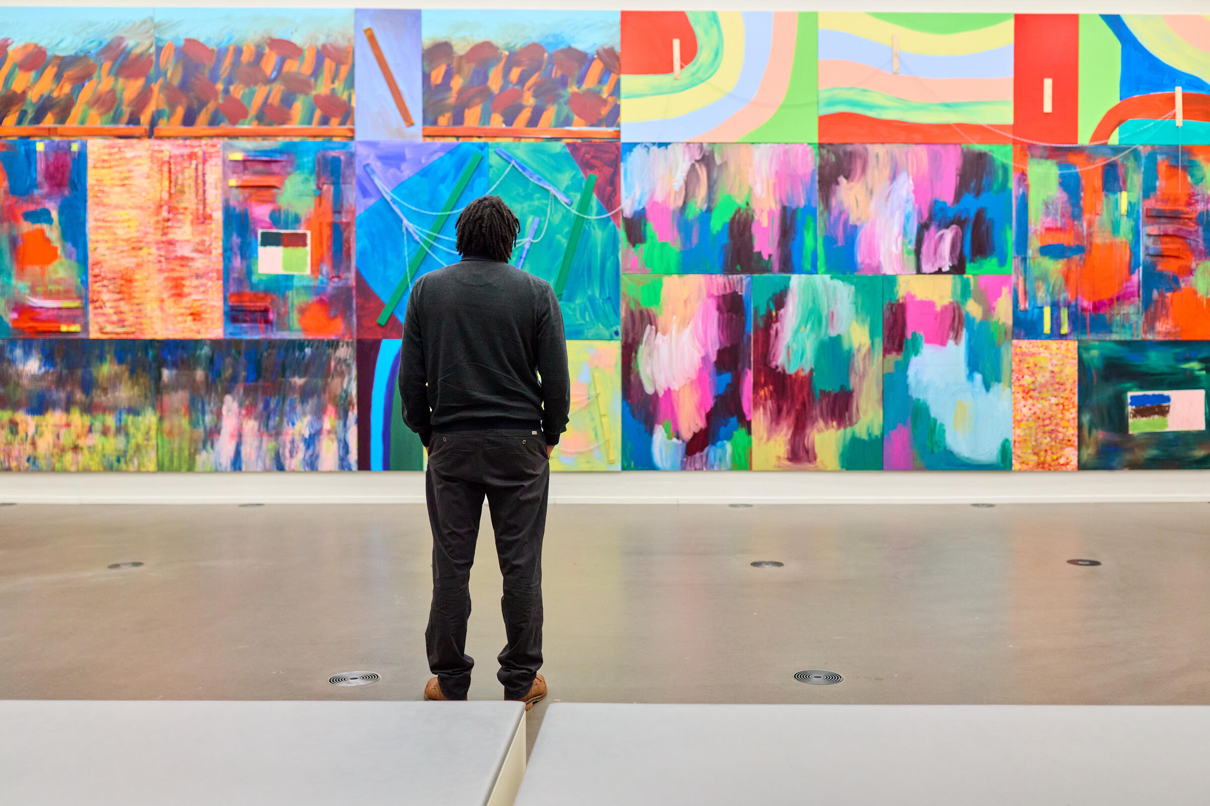 Een man in zwart staat voor een kleurrijk schilderij dat de hele muur vult, met abstracte vormen en felle kleuren.