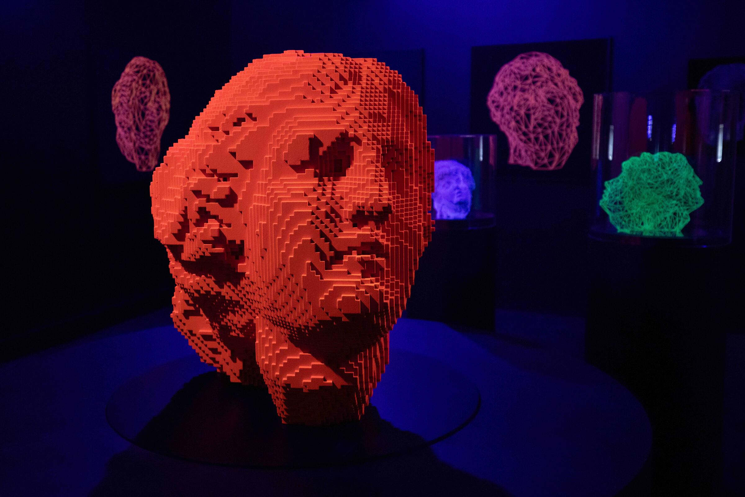 Eine Skulptur, die den Kopf einer Frau darstellt, deren Frisur und Gesichtsz&uuml;ge an die Statuen aus der Antike erinnern. Nur ist diese Skulptur komplett pixelig und knallrot. Hinter der Skulptur h&auml;ngen in einem dunkelvioletten Raum Bildschirme.