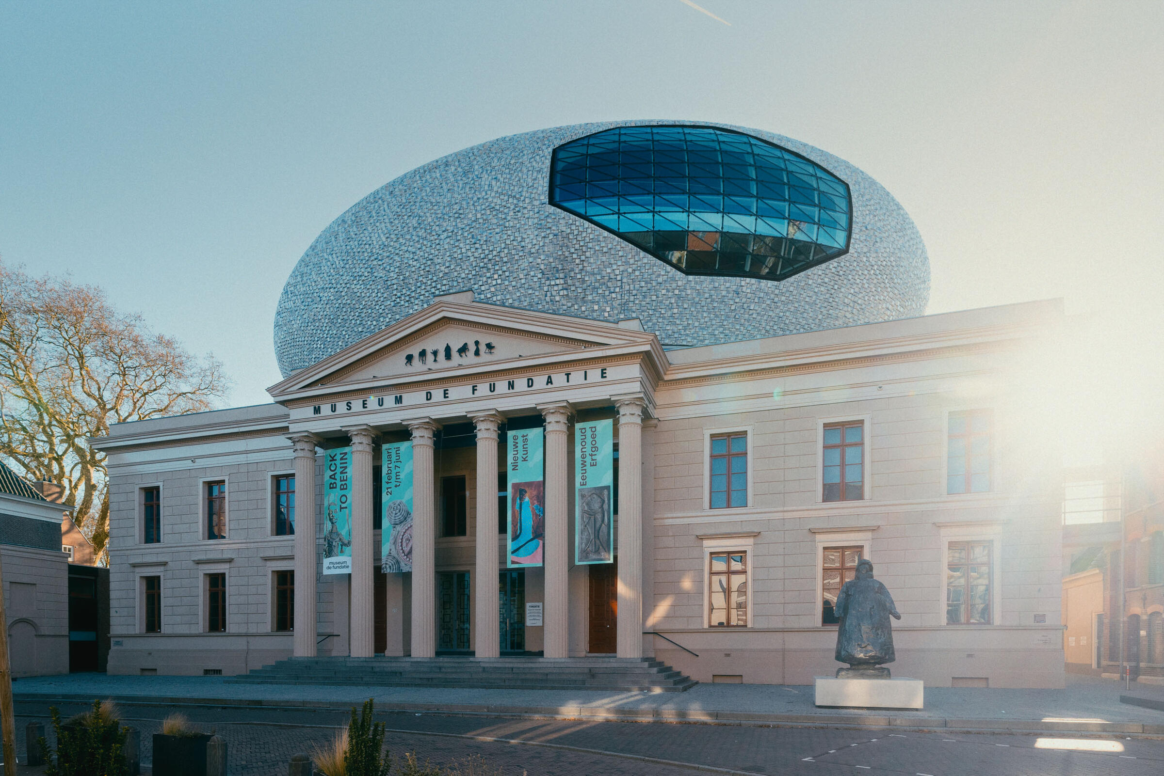 Een vooraangezicht van Museum de Fundatie met de pilaren en het ovale dak goed zichtbaar in het zonlicht.