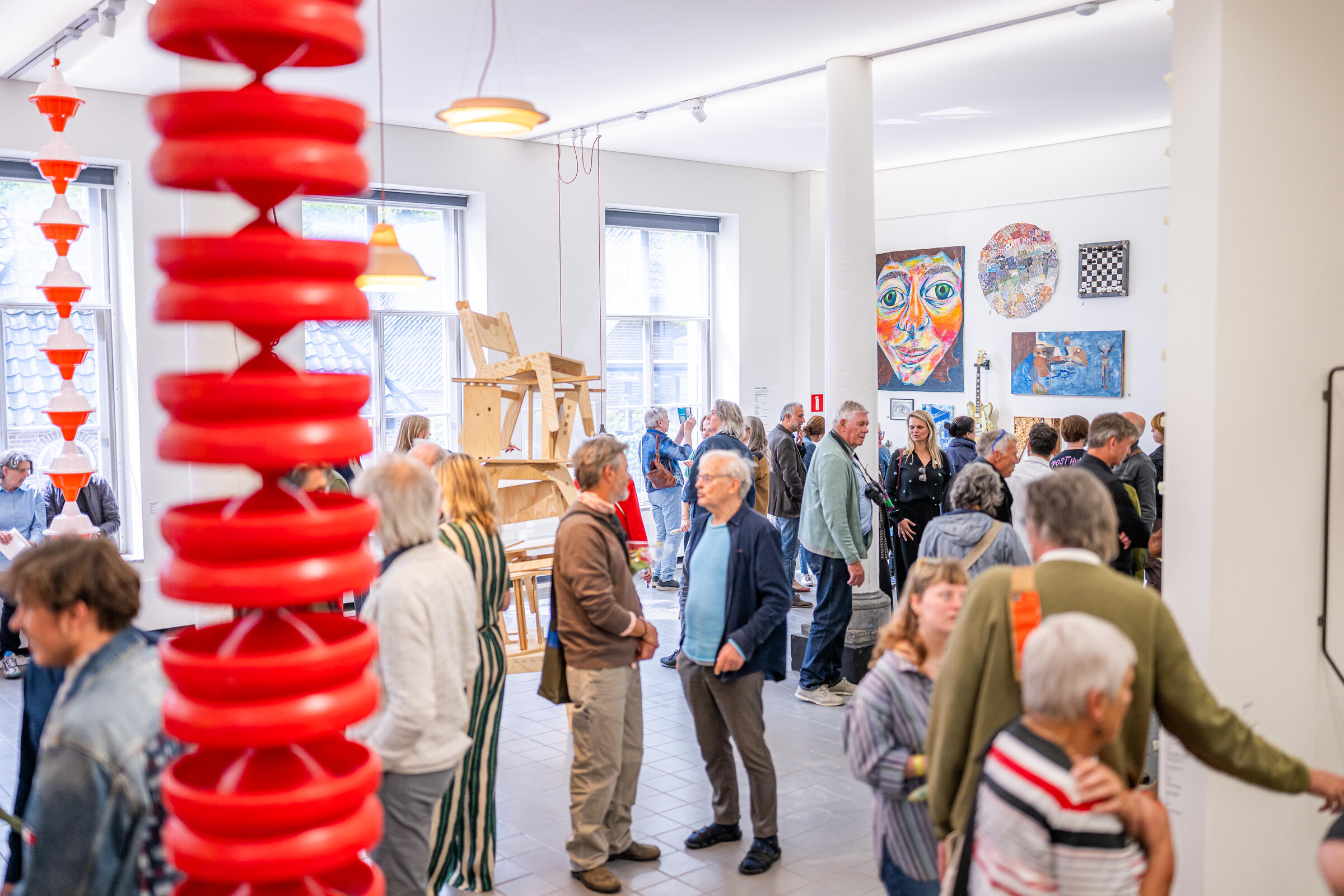 Tientallen bezoekers staan in de expositieruimte. Aan de voorkant van de foto hangt een rood kunstwerk van de plafond. 