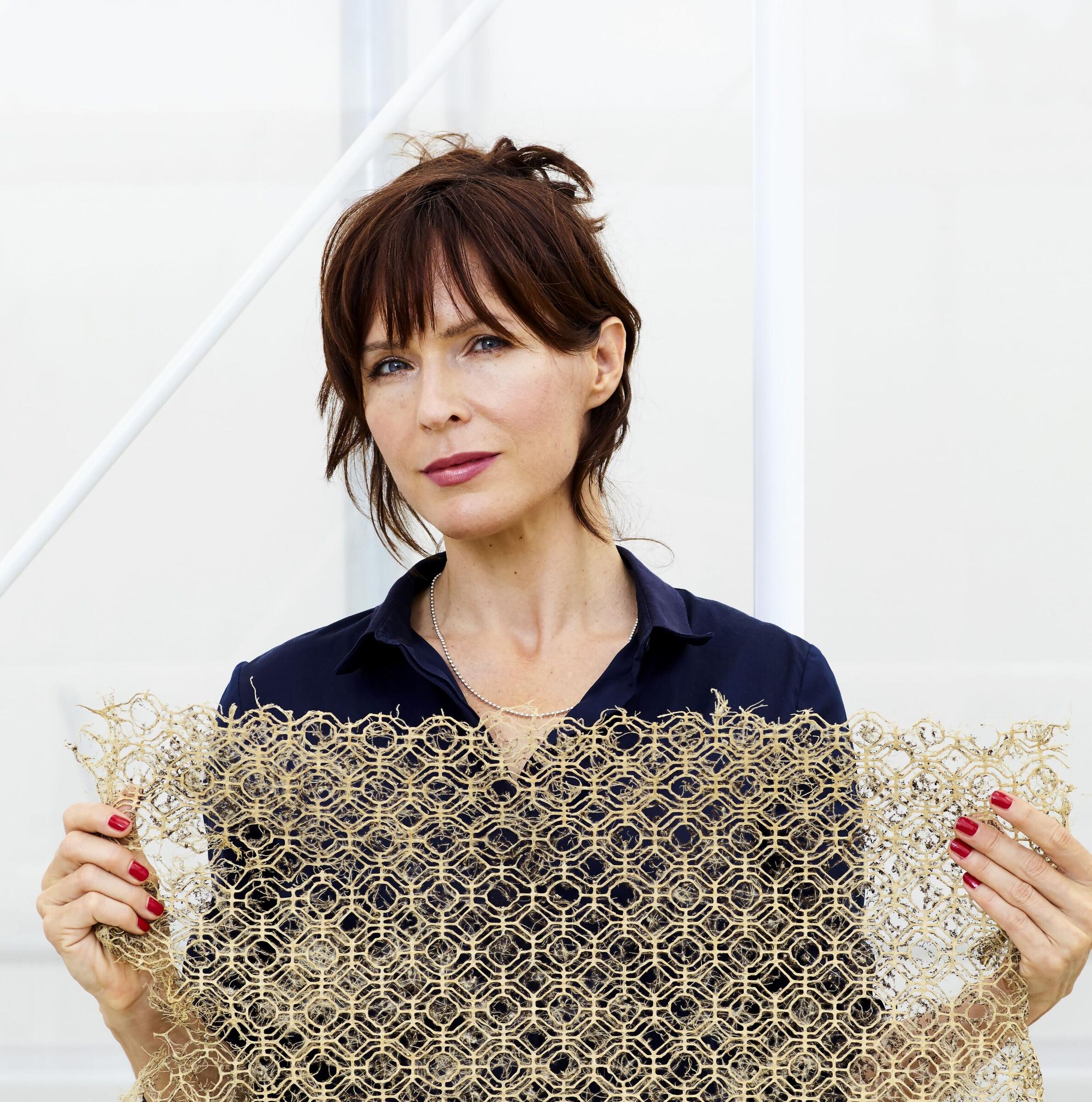 Diana Scherer houdt een van haar kunstwerken, een combinatie van fragiel textiel en wortels omhoog. Ze kijkt in de camera.