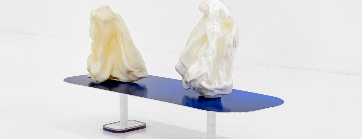 Twee witte sculpturen die lijken op proppen papier of zijde, rustend op een blauwe tafel.