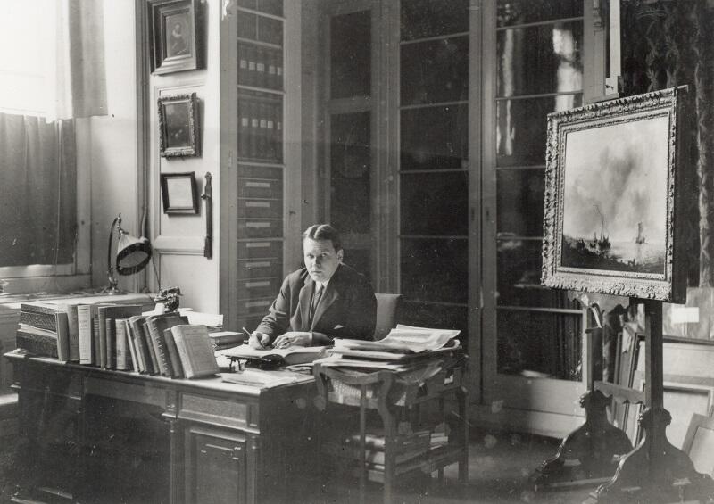 Dirk Hannema zit aan een bureau als volwassen man van rond de 35. Hij is omringd met kunst en boeken.