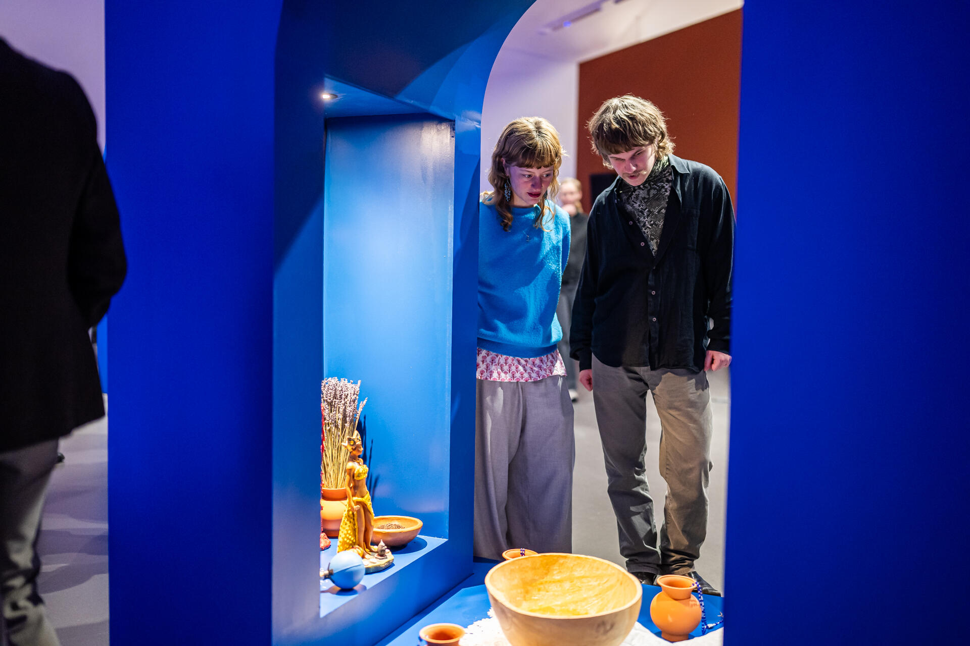 Twee jonge mensen kijken naar een blauwe boog. In de boog staat een houten kom, aardewerken schalen en beelden van vrouwen en vruchtbaarheid.
