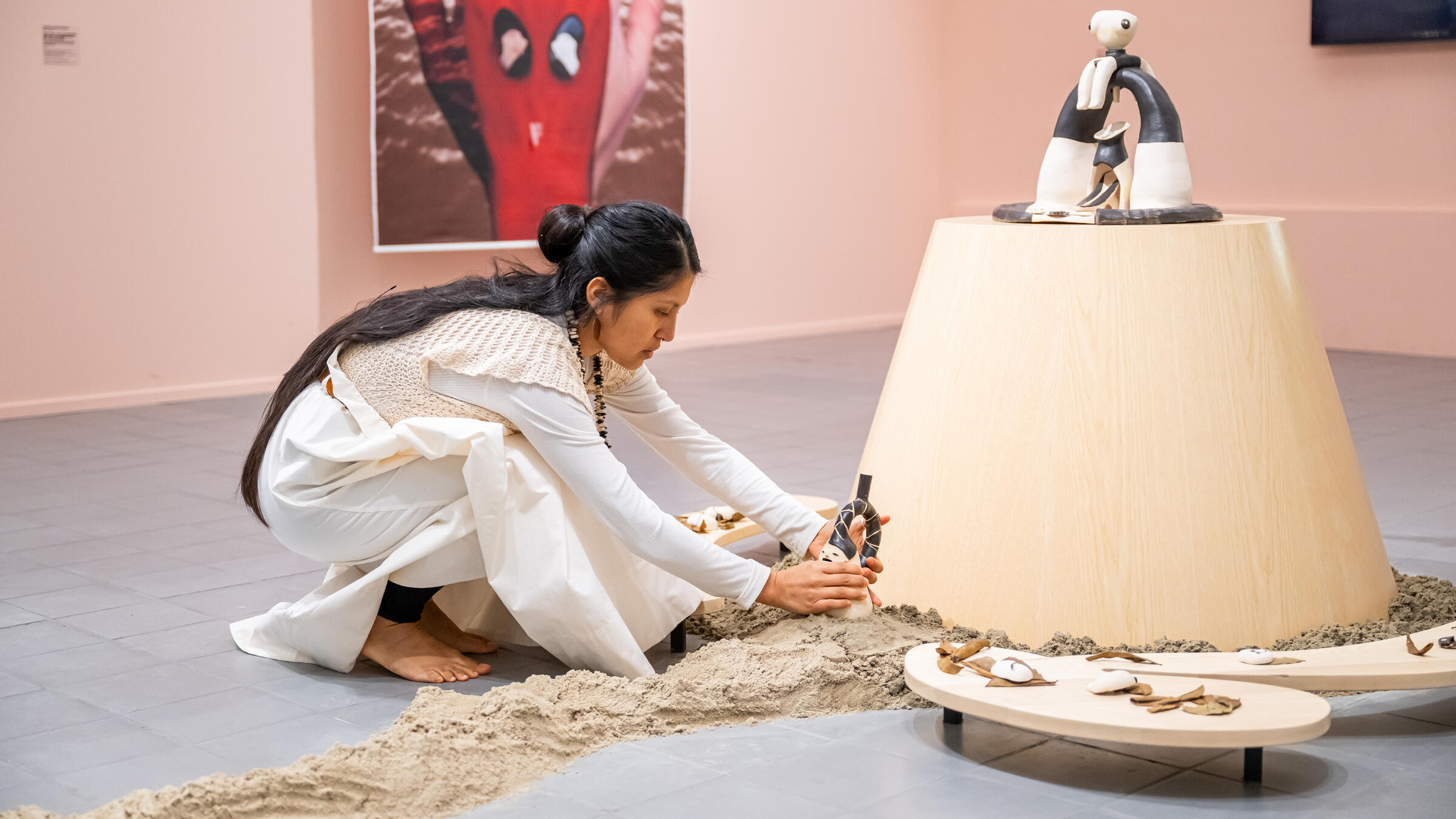 Cristina Flores Pescorán bij haar eigen kunstwerk. Het kunstwerk bestaat uit verschillende materialen zoals zand, hout en beeldhouw kunstwerken. De kleuren zijn licht en naturel.