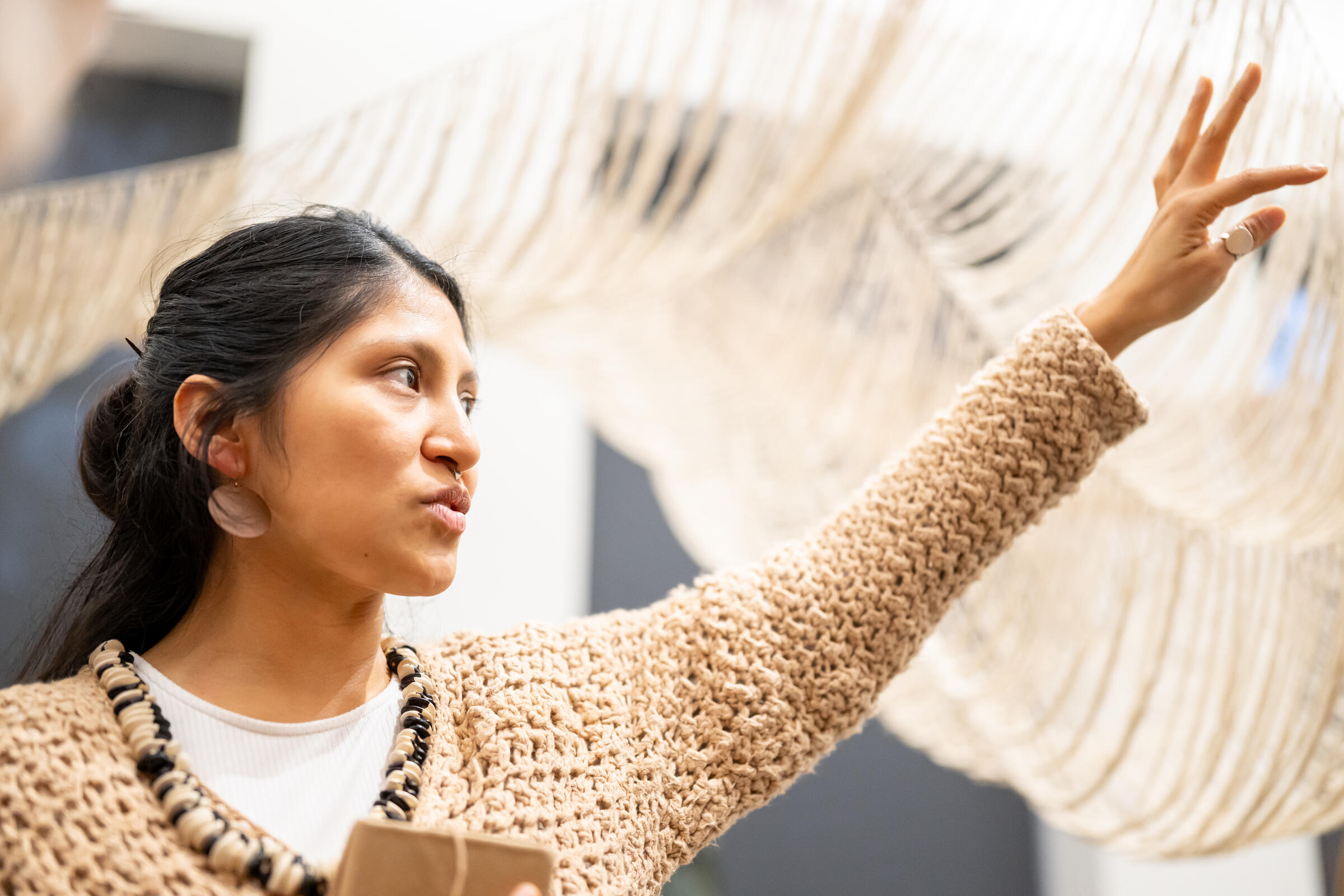 een close-up foto van Cristina Flores Pescorán, ze houdt haar arm omhoog. In de achtergrond zie je haar zandgekleurde geweven werk