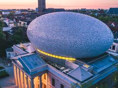 An aerial view of Museum De Fundatie in Zwolle. It is dusk and the lights beneath the columns cast a warm glow. The sky is pale pink.
