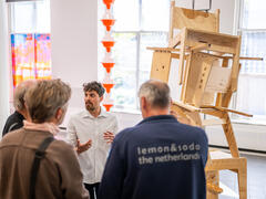 A young artist stands by an artwork of stacked wooden chairs. He addresses a group of visitors.