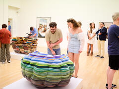 Bezoekers bekijken kunstwerken in een lichte museumzaal, waaronder een grote, kleurrijke, gelaagde sculptuur op een sokkel en een stapel boeken op de achtergrond.